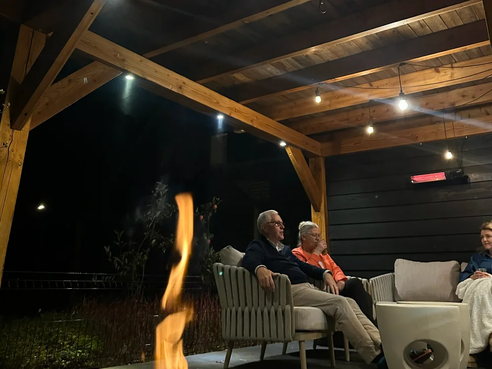 Warme buitenverlichting onder een houten overkapping met terras, vuur en gezellige zithoek in de avond
