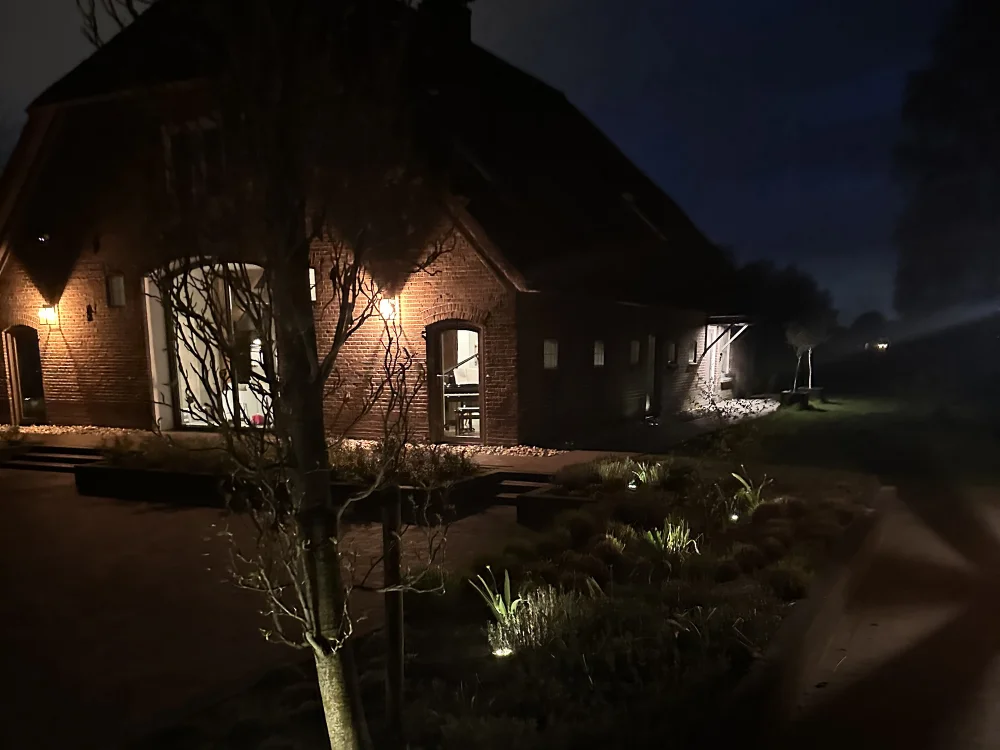 Sfeervolle tuinverlichting bij een landelijke woning met verlichte gevel, planten en looppad in de avond